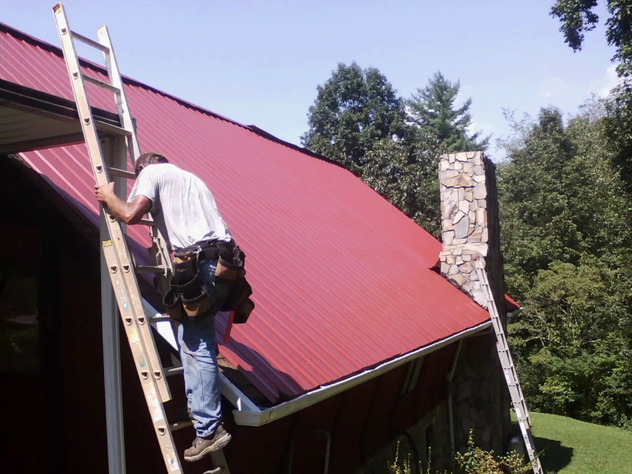 Roofer on a ladder beside a red metal roof and stone chimney — Soffit & Fascia Repair by Roofing Brothers in Mooreville